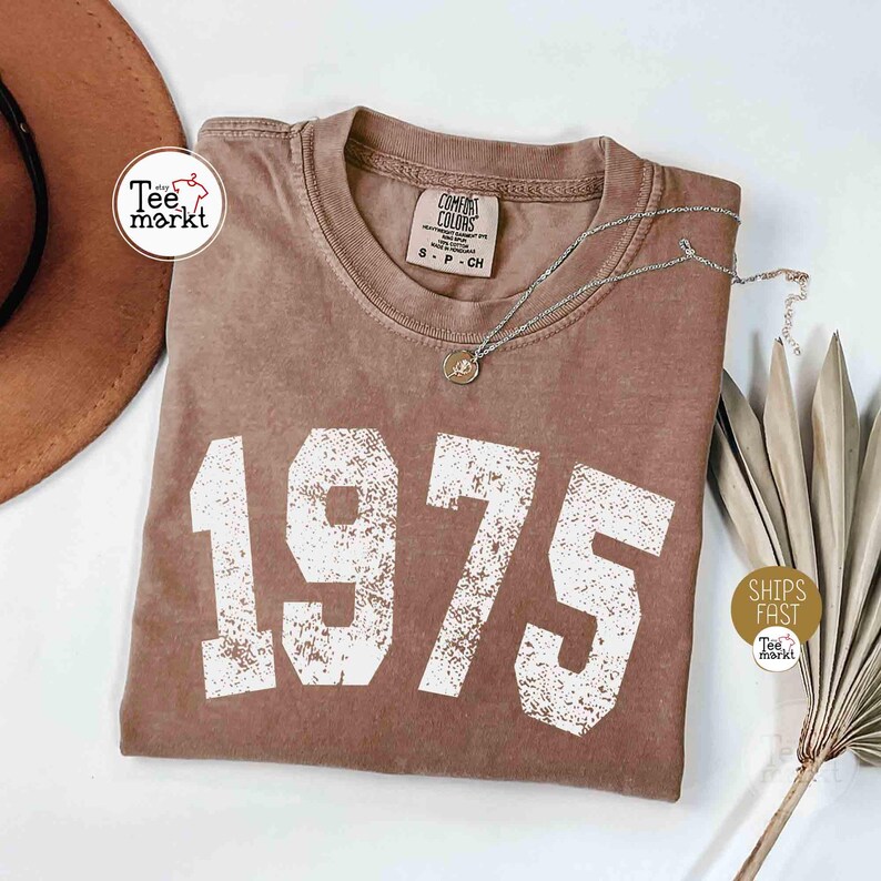 May include: A brown t-shirt with the year "1975" printed in large, white, distressed lettering. A silver necklace with a pendant rests on the shirt. The shirt is from the "Comfort Colors" brand and has a small tag. A brown hat is visible in the background.