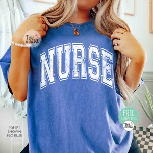May include: A blue t-shirt with the word "NURSE" in large, distressed white lettering. The shirt is a solid color and has a crew neck. The image also shows a bracelet and a necklace.