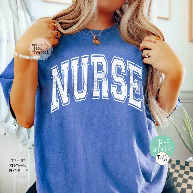 May include: A blue t-shirt with the word "NURSE" in large, distressed white lettering. The shirt is a solid color and has a crew neck. The image also shows a bracelet and a necklace.