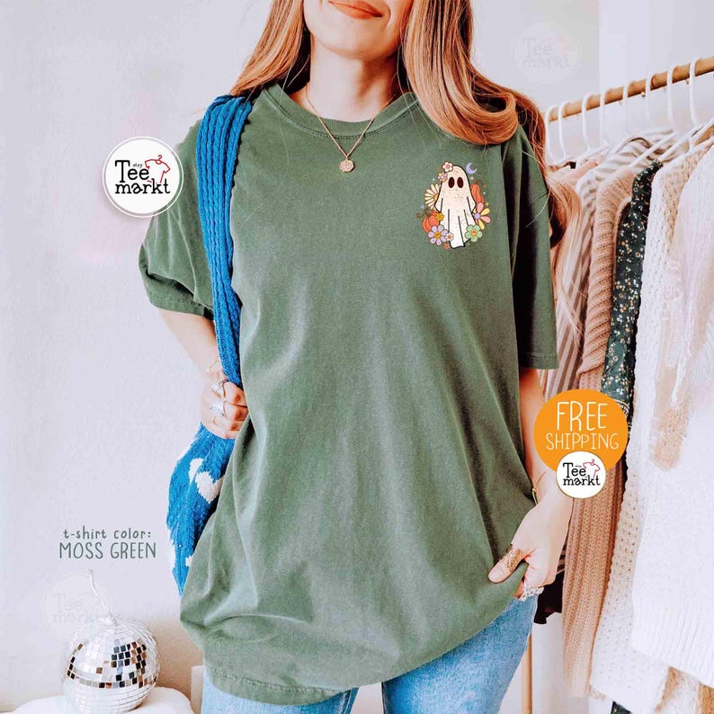 以下が含まれることがあります： モスグリーンのTシャツ。漫画のゴーストとカラフルな花のデザイン。Tシャツは半袖でゆったりとしたフィット感です。Tシャツのテキストは「t-shirt color: MOSS GREEN」と書かれています。