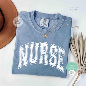 May include: A blue jean colored t-shirt with the word "NURSE" in large white block letters. The shirt has a crew neck and short sleeves. A gold necklace with a pendant is draped across the shirt. The shirt is from Tee Markt.
