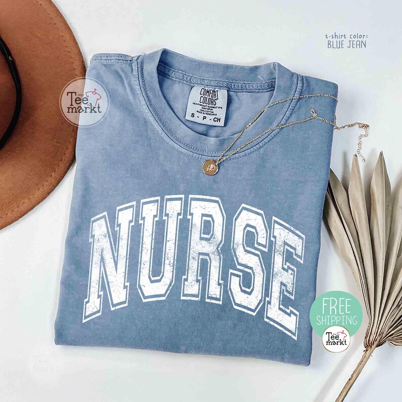 May include: A blue jean colored t-shirt with the word "NURSE" in large white block letters. The shirt has a crew neck and short sleeves. A gold necklace with a pendant is draped across the shirt. The shirt is from Tee Markt.