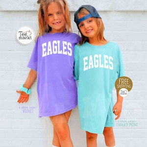 May include: Two young girls wearing oversized t-shirts with the word "EAGLES" in white block letters. One shirt is violet, the other is chalky mint. The shirts are labeled with the colors. One girl wears a blue cap.