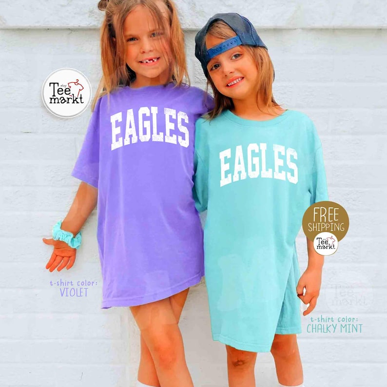 May include: Two young girls wearing oversized t-shirts with the word "EAGLES" in white block letters. One shirt is violet, the other is chalky mint. The shirts are labeled with the colors. One girl wears a blue cap.