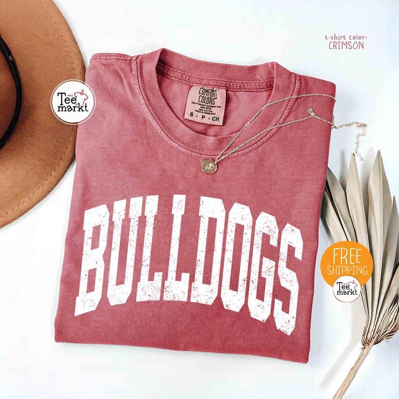 May include: Crimson-colored t-shirt with the word "BULLDOGS" in white block letters. A gold necklace with a pendant rests on the shirt. The shirt has a vintage, washed look. The text "Tee markt" is visible.