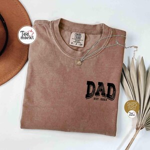 May include: A brown Comfort Colors t-shirt with the word "DAD" in a black, vintage-style font, and "EST. 2023" below it. A silver necklace with a pendant rests on the shirt. The shirt is on a white surface.