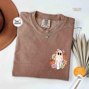 以下が含まれることがあります： エスプレッソ色のTシャツ。花、カボチャ、三日月で囲まれた幽霊のグラフィックが特徴です。Tシャツはクルーネックと半袖です。ペンダント付きのシルバーネックレスがTシャツの上にあります。「Tee markt」の文字が見えます。
