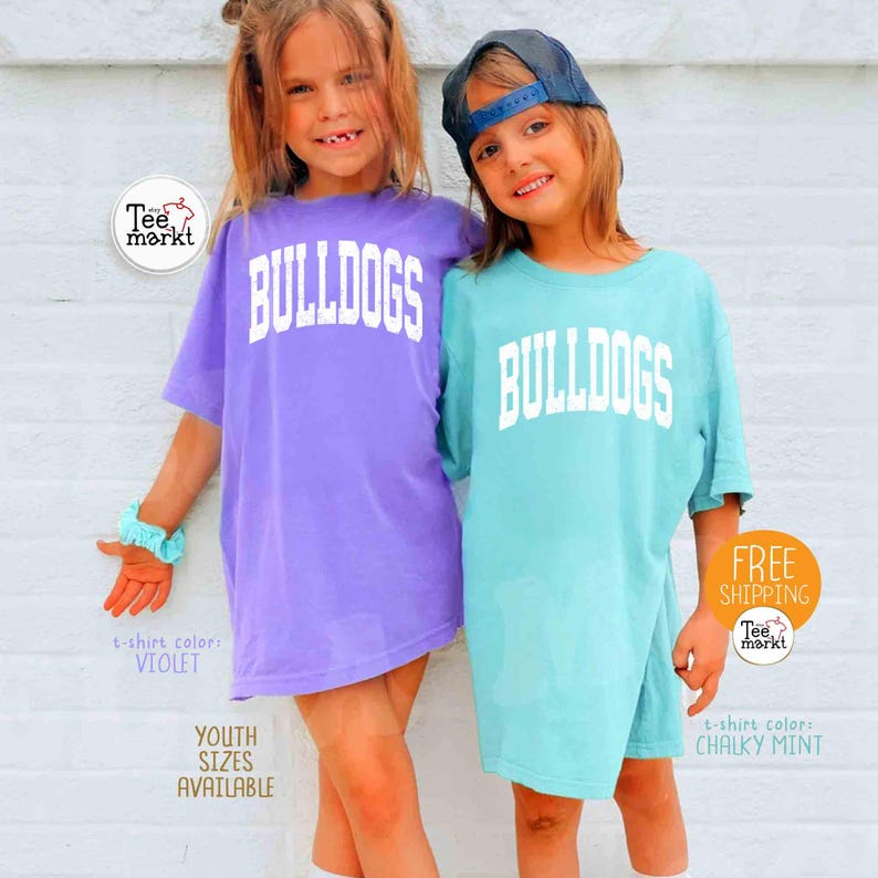 May include: Two children wearing oversized t-shirts with the word "BULLDOGS" in white block letters. One shirt is violet, the other is chalky mint. The image includes the text "YOUTH SIZES AVAILABLE".