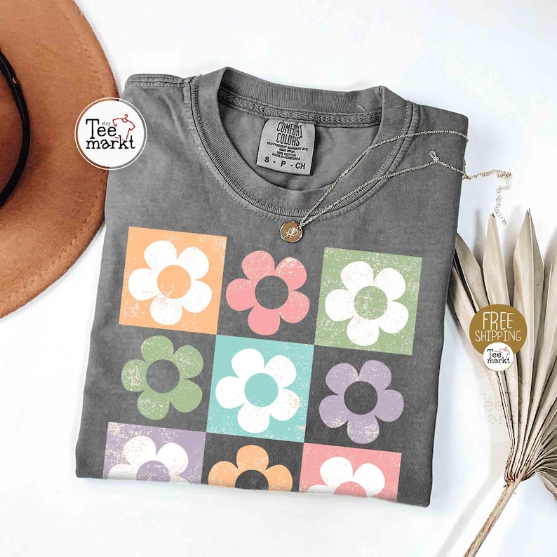 May include: Gray t-shirt with a colorful floral design. The shirt features nine squares, each with a different colored flower. A silver necklace with a small pendant rests on the shirt. The shirt is from Comfort Colors.