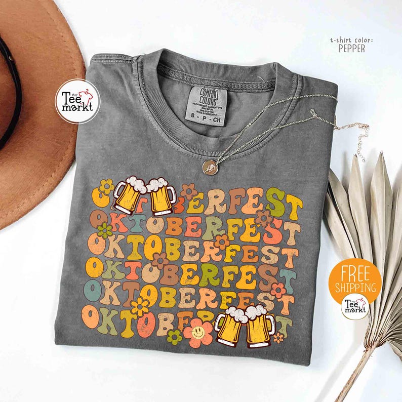 K&ouml;nnte beinhalten: Graues T-Shirt mit dem Wort "OKTOBERFEST" in einer bunten Retro-Schriftart. Das Design umfasst Bierkr&uuml;ge und Blumen. Marke Comfort Colors, Gr&ouml;&szlig;e S-P-CH. T-Shirt-Farbe: Pfeffer.