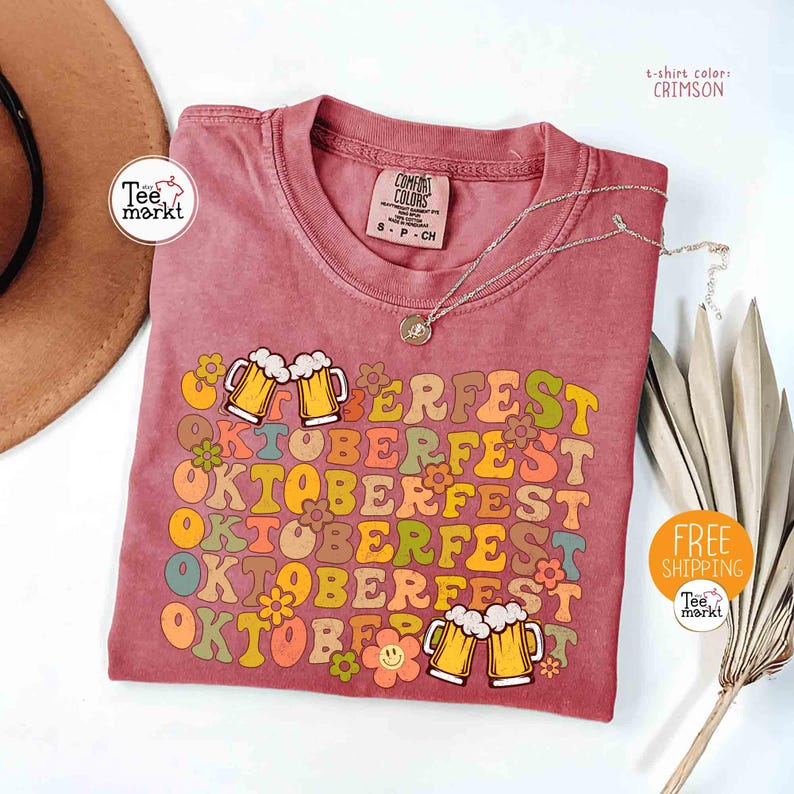 K&ouml;nnte beinhalten: Karmesinrotes T-Shirt mit dem Wort "OKTOBERFEST" in Retro-Schrift, verziert mit Bierkr&uuml;gen und Blumen. Das Shirt hat ein Comfort Colors-Etikett und eine silberne Halskette. Gr&ouml;&szlig;e S-P-CH.