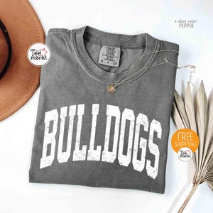 May include: A gray t-shirt with the word "BULLDOGS" in large, distressed white letters. The shirt has a crew neck and short sleeves. A gold pendant necklace rests on the shirt. The t-shirt color is Pepper.