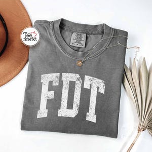 May include: A gray Comfort Colors t-shirt with the white letters "FDT" in a distressed font. A silver necklace with a pendant rests on the shirt. A brown hat and dried plant are in the background.