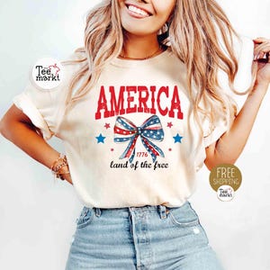 May include: Cream-colored t-shirt with the word "AMERICA" in red, a patriotic bow graphic, and the text "1776 land of the free." The design includes red, white, and blue stars and a bow, celebrating American independence.