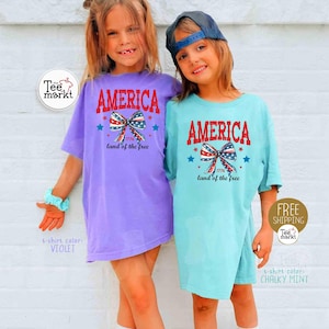 May include: Two young children wearing matching t-shirts. The shirts are violet and chalky mint, each with the word "AMERICA" in red, a bow with red, white, and blue accents, and the text "1776 land of the free".
