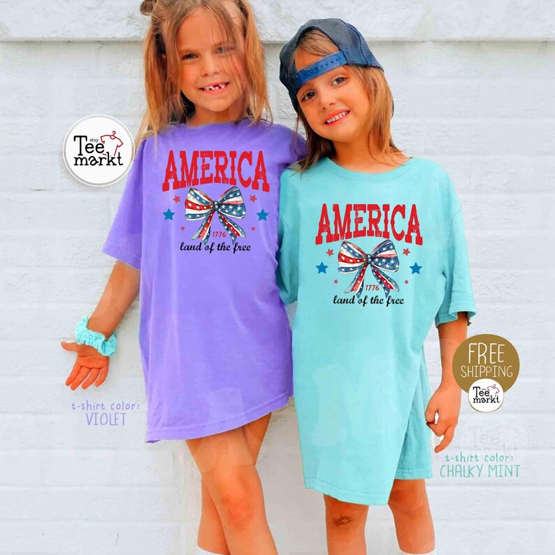May include: Two young children wearing matching t-shirts. The shirts are violet and chalky mint, each with the word "AMERICA" in red, a bow with red, white, and blue accents, and the text "1776 land of the free".