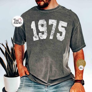 May include: Dark gray t-shirt with the number "1975" printed in distressed white lettering. The shirt has a vintage, worn look. The image also includes a plant in a white pot and a person wearing the shirt.