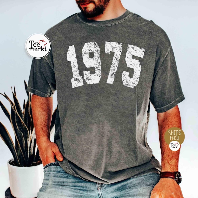 May include: Dark gray t-shirt with the number "1975" printed in distressed white lettering. The shirt has a vintage, worn look. The image also includes a plant in a white pot and a person wearing the shirt.