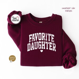 May include: A maroon crewneck sweatshirt featuring the white distressed text FAVORITE DAUGHTER arched across the chest with a small heart icon centred below the lettering. The garment has long sleeves and a ribbed collar, cuffs, and hem.