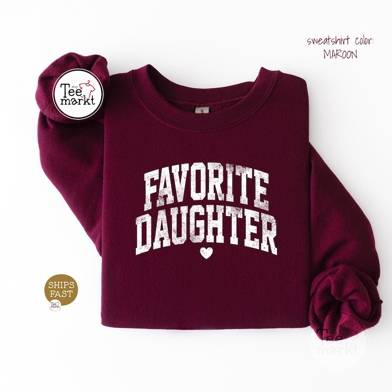May include: A maroon crewneck sweatshirt featuring the white distressed text FAVORITE DAUGHTER arched across the chest with a small heart icon centred below the lettering. The garment has long sleeves and a ribbed collar, cuffs, and hem.
