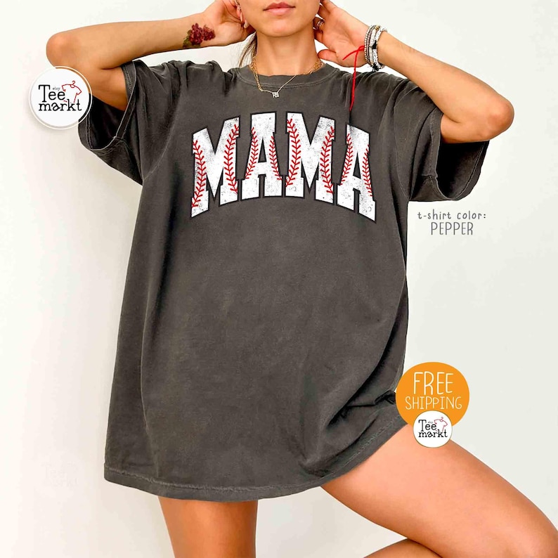 May include: Pepper-coloured oversized t-shirt with the word "MAMA" in white block letters, featuring red baseball stitching. The shirt has a vintage, worn look.
