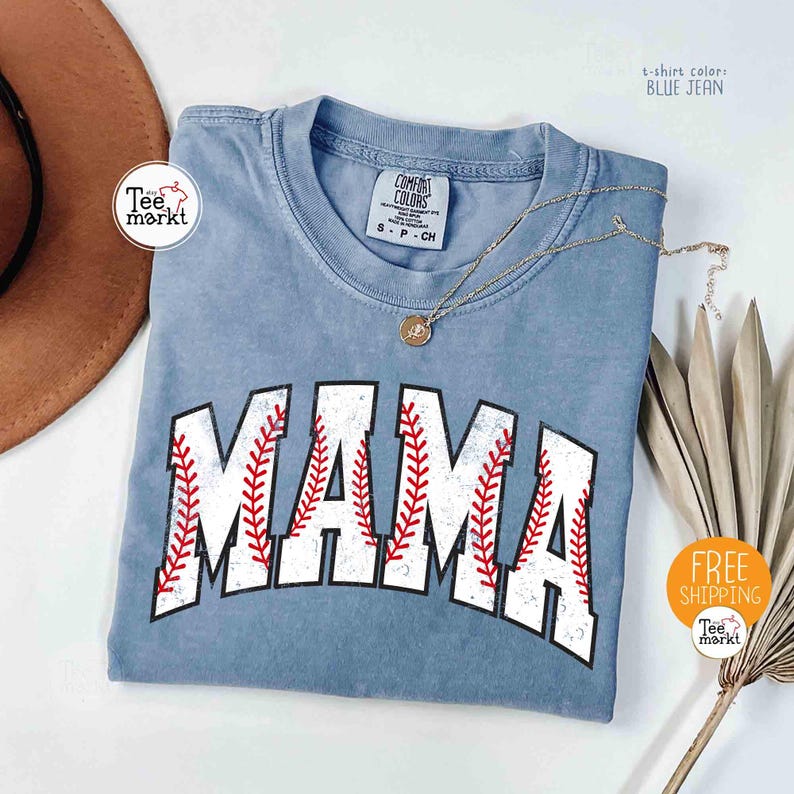 May include: A blue jean coloured t-shirt with the word "MAMA" in white block letters with red baseball stitching. A gold necklace with a pendant rests on the shirt. The shirt has a crew neck and short sleeves. The shirt is from Comfort Colors.