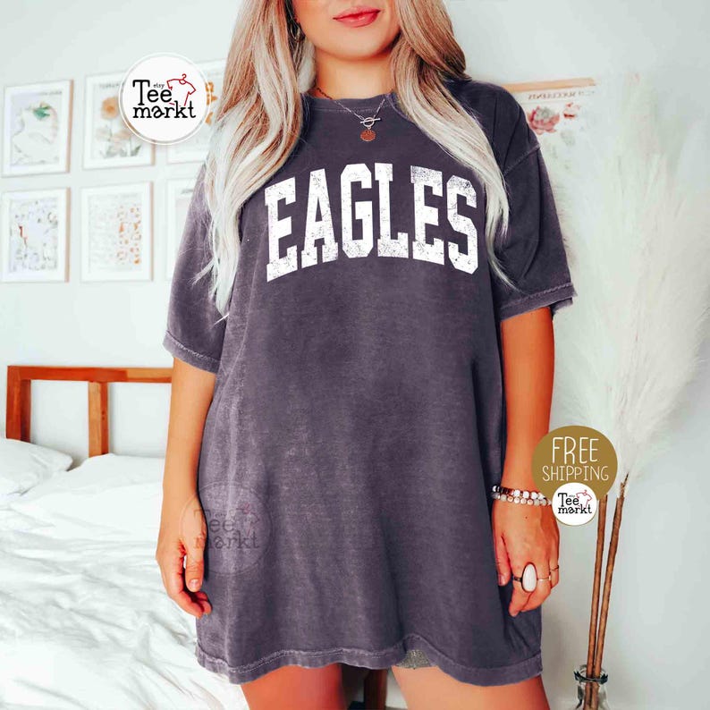 May include: A dark gray t-shirt with the word "EAGLES" in large, white, distressed lettering across the chest. The shirt has short sleeves and a relaxed fit. The image also includes a "Free Shipping" sticker.