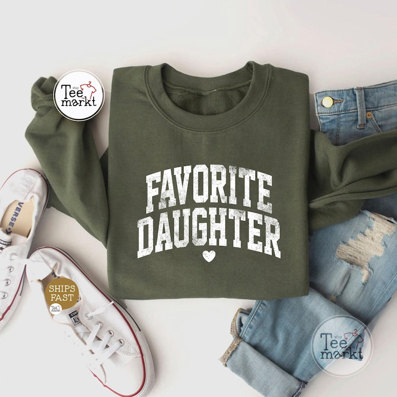 May include: An olive green crewneck sweatshirt featuring the white distressed text "FAVORITE DAUGHTER" in a bold, arched collegiate font with a small white heart icon centred below the lettering, paired with blue denim jeans and white trainers.