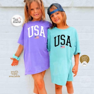 May include: Two children wearing oversized t-shirts with "USA" printed in large letters above a small American flag. One shirt is purple, the other is light blue. The shirts are part of a youth collection.