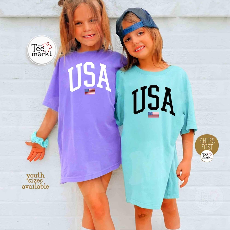 May include: Two children wearing oversized t-shirts with "USA" printed in large letters above a small American flag. One shirt is purple, the other is light blue. The shirts are part of a youth collection.