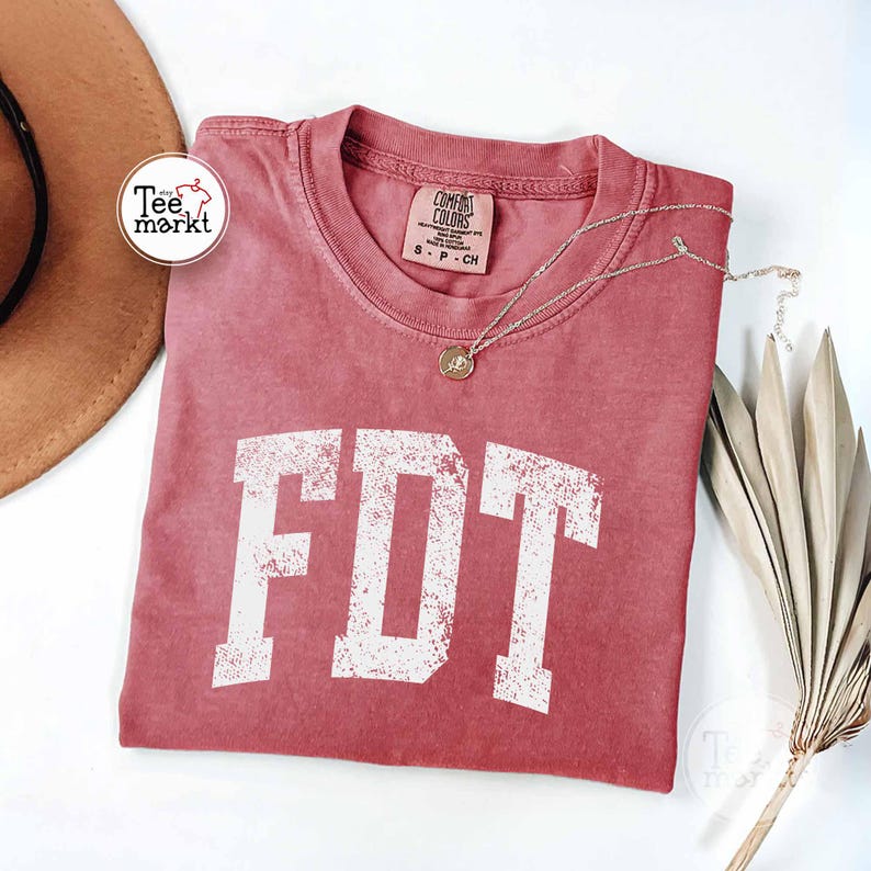 May include: A red-orange t-shirt with the letters "FDT" in distressed white lettering. The shirt has a crew neck and short sleeves. A gold necklace with a pendant is draped across the shirt. A brown hat and dried plant are in the background.