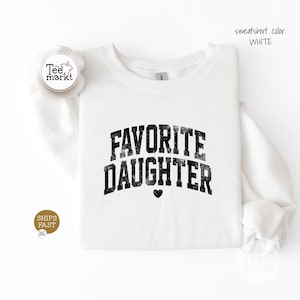 May include: A white crewneck sweatshirt featuring the black distressed text FAVORITE DAUGHTER in an arched collegiate font with a small black heart icon centred below the lettering. The text sweatshirt colour: WHITE is visible in the upper right corner.