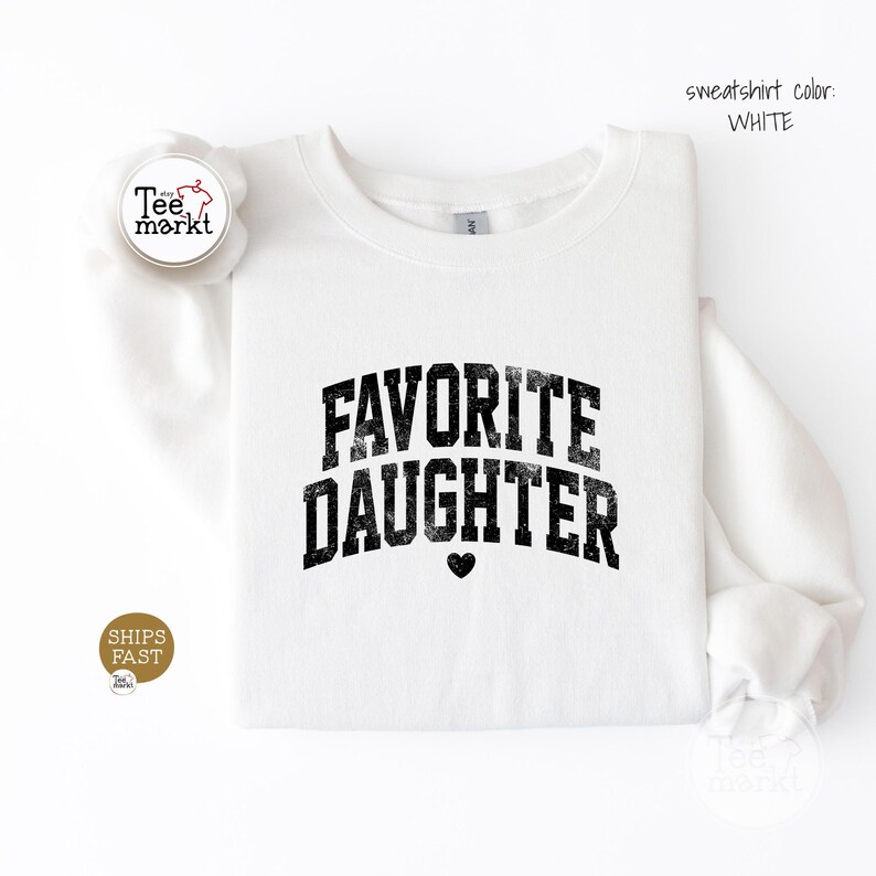 May include: A white crewneck sweatshirt featuring the black distressed text FAVORITE DAUGHTER in an arched collegiate font with a small black heart icon centred below the lettering. The text sweatshirt colour: WHITE is visible in the upper right corner.