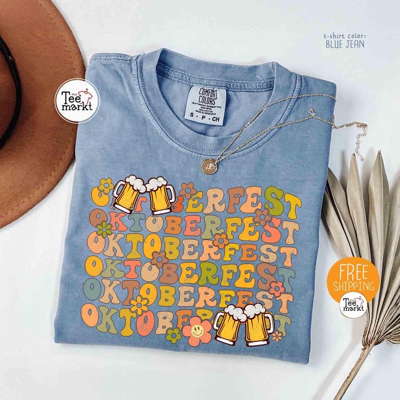 K&ouml;nnte beinhalten: Blaues Jeans-T-Shirt mit dem Wort "OKTOBERFEST" in Retro-Schrift, verziert mit Bierkr&uuml;gen und Blumen. Eine goldene Halskette und ein brauner Hut sind ebenfalls sichtbar. Das Shirt ist mit "Comfort Colors" gekennzeichnet.