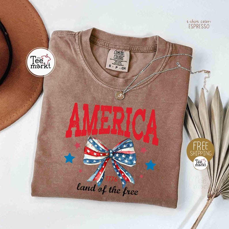 May include: Espresso-colored t-shirt with the word "AMERICA" in red block letters. A patriotic bow graphic with red, white, and blue stars and stripes is below the word. The text "land of the free" is printed below the bow.