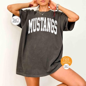 May include: Dark gray t-shirt with the word "MUSTANGS" in large white block letters. The shirt has a vintage, worn look. The shirt is a crew neck and has short sleeves.