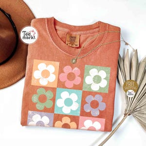 May include: Orange Comfort Colors t-shirt with a floral design. The design features nine colorful flowers in square blocks. A gold necklace with a pendant is on the shirt. A brown hat and a dried plant are in the background.