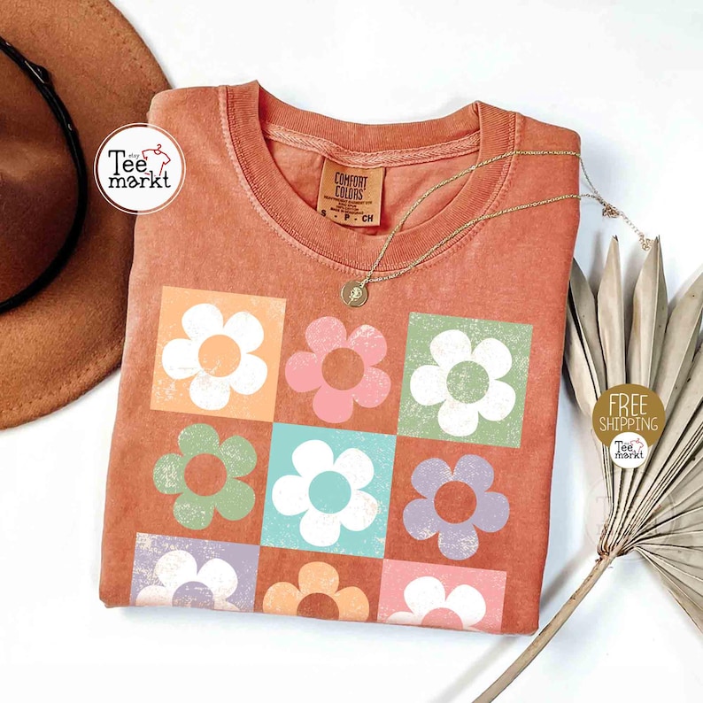 May include: Orange Comfort Colors t-shirt with a floral design. The design features nine colorful flowers in square blocks. A gold necklace with a pendant is on the shirt. A brown hat and a dried plant are in the background.