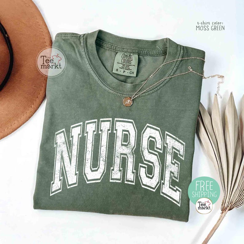 May include: Moss green t-shirt with the word "NURSE" in large, distressed white letters. A gold necklace with a pendant rests on the shirt. The shirt is folded, with a brown hat and dried plant in the background.