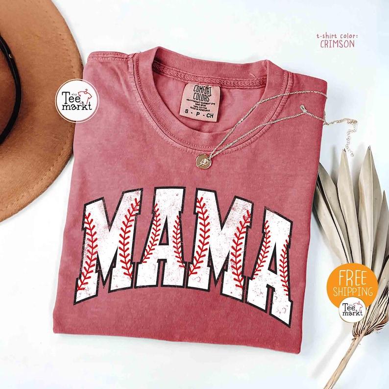 May include: Crimson t-shirt featuring the word "MAMA" in a baseball-style font. The letters are white with red stitching. A gold necklace and a decorative plant are also visible.