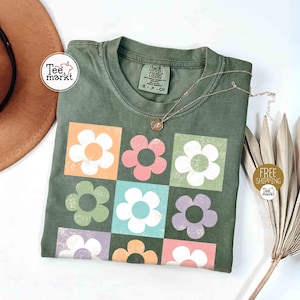 May include: Olive green t-shirt with a floral design. The design features nine colorful flowers in square blocks. A gold necklace with a pendant rests on the shirt. The shirt has a tag that says "COMFORT COLORS".