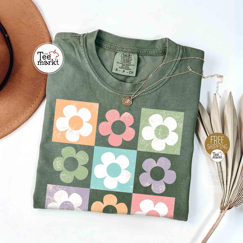 May include: Olive green t-shirt with a floral design. The design features nine colorful flowers in square blocks. A gold necklace with a pendant rests on the shirt. The shirt has a tag that says "COMFORT COLORS".