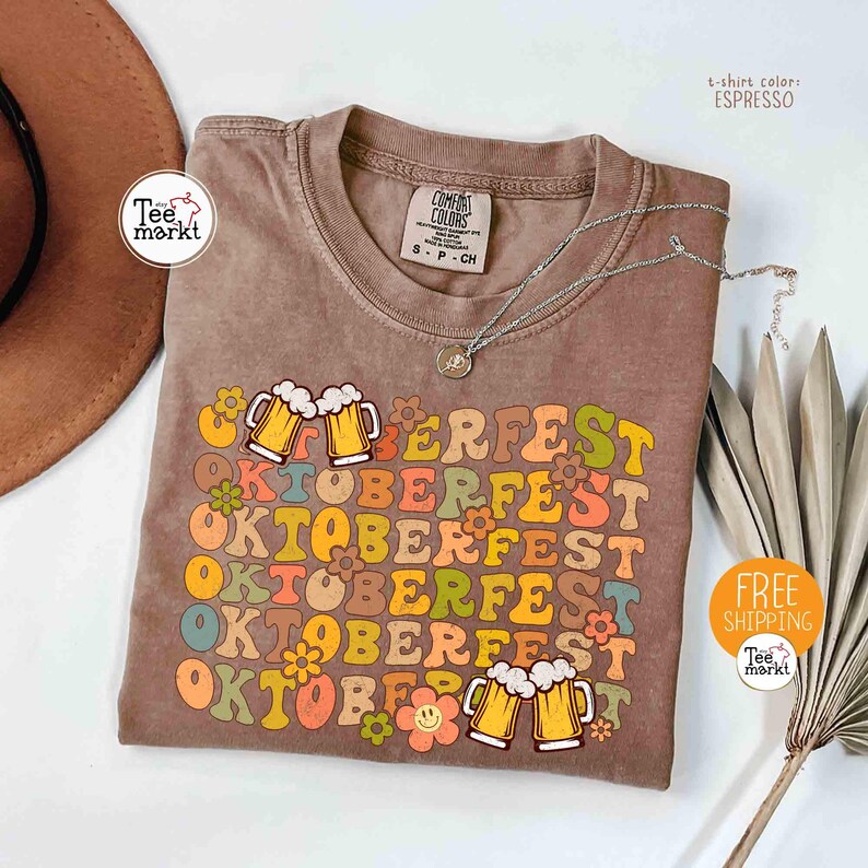 K&ouml;nnte beinhalten: Espressofarbenes T-Shirt mit einem Retro-Design "Oktoberfest". Das Design zeigt Bierkr&uuml;ge, Blumen und das Wort "Oktoberfest" in verschiedenen Farben. Eine silberne Halskette liegt &uuml;ber dem Shirt.