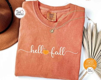 May include: Orange-colored t-shirt with the words "hello fall" in white script, featuring a small pumpkin graphic. The shirt has a crew neck and a Comfort Colors tag. A gold necklace with a pendant is draped across the shirt. A brown hat is visible in the background.