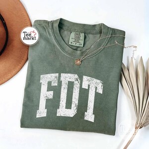 May include: An olive green t-shirt with the white letters "FDT" in a distressed, collegiate font. A gold-colored necklace with a pendant rests on the shirt. The shirt has a crew neck and a tag that reads "Comfort Colors".