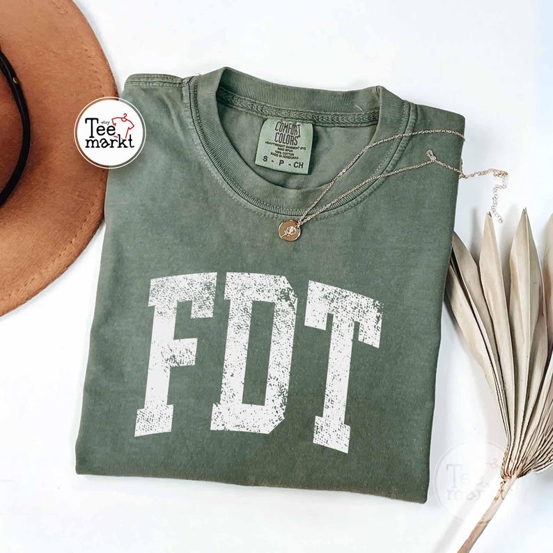 May include: An olive green t-shirt with the white letters "FDT" in a distressed, collegiate font. A gold-colored necklace with a pendant rests on the shirt. The shirt has a crew neck and a tag that reads "Comfort Colors".
