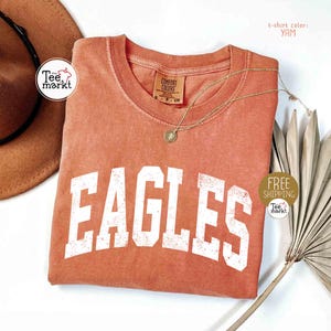 May include: A coral-colored t-shirt with the word "EAGLES" in large, distressed white letters. The shirt has a crew neck and a small tag. A gold necklace with a pendant is draped across the shirt. The t-shirt color is "YAM".