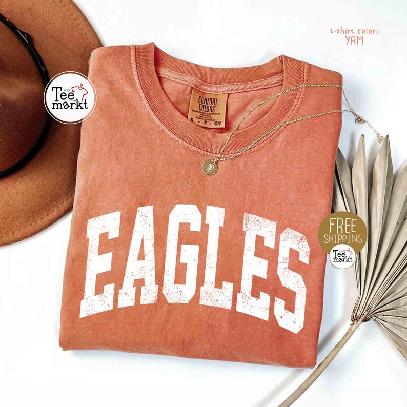 May include: A coral-colored t-shirt with the word "EAGLES" in large, distressed white letters. The shirt has a crew neck and a small tag. A gold necklace with a pendant is draped across the shirt. The t-shirt color is "YAM".