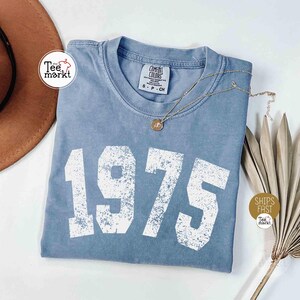 May include: A blue t-shirt with the year "1975" printed in large white block letters. The shirt has a crew neck and short sleeves. A gold necklace with a pendant rests on the shirt. A brown hat and dried plant are in the background.