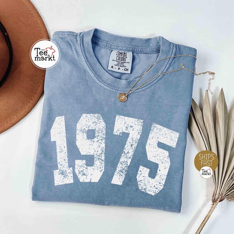 May include: A blue t-shirt with the year "1975" printed in large white block letters. The shirt has a crew neck and short sleeves. A gold necklace with a pendant rests on the shirt. A brown hat and dried plant are in the background.